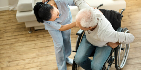 Eine junge Pflegekraft unterstützt einen alten Mann beim Aufstehen aus einem Rollstuhl der sich dabei mit dem Arm an ihren Schultern abstützt