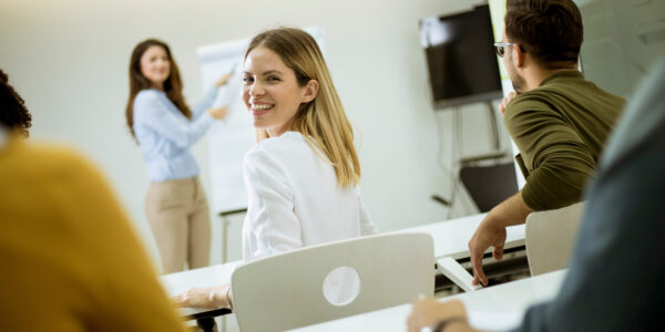 Eine Gruppe junger Menschen wird gerade von einer Frau unterrichtet. Eine junge Auszubildende dreht sich dabei um und schaut selbstbewusst lachend in die Kamera