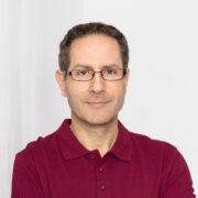 Andreas Schumann, Inhaber des APS-Instituts lächelnd in einem rostroten Polo-Shirt an eine weiße Wand gelehnt